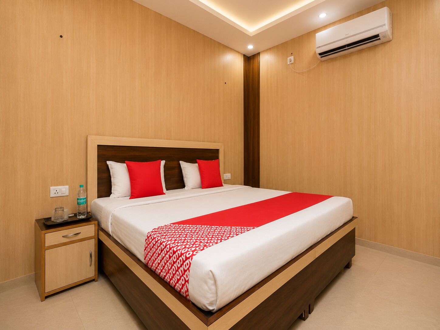 Una habitación de hotel con una cama de madera, una mesita de noche, una botella de agua y un aire acondicionado.