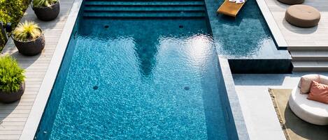 A rectangular swimming pool with a built-in seating area, surrounded by potted plants and outdoor furniture.