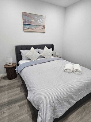Ein ordentlich bezogenes Bett mit gestreifter Bettwäsche, ein grauer möbelbezogener Kopfteil und ein kleiner Nachttisch mit einer Lampe und einem runden Holzgegenstand.