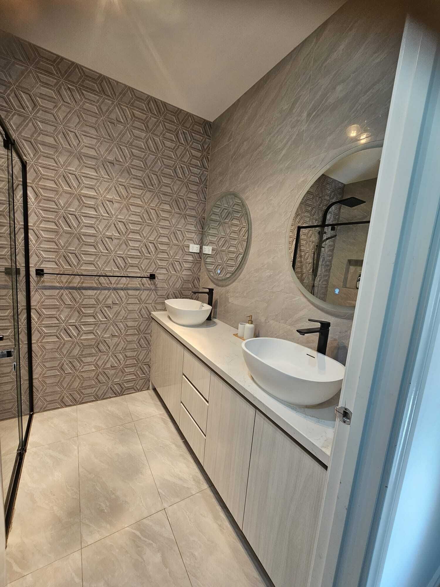 Een moderne badkamer met een wand in geometrisch patroon, een ronde spiegel, twee wasbakken en een douchecabine met glazen wanden.