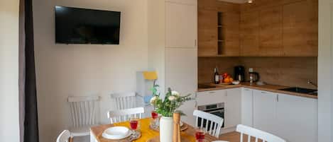 A dining area with a wooden table set for four, white chairs, a TV mounted on the wall, and a kitchen with wooden cabinets and appliances.