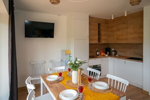 Comedor con mesa de madera dispuesta para cuatro, sillas blancas, televisor montado en la pared y cocina con armarios y electrodomésticos de madera.