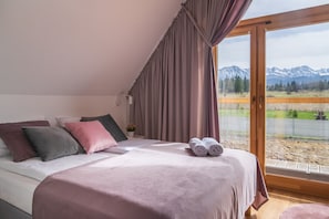 A bedroom with a bed, pillows, a small table, and a window with a mountain view. - Miodowe Domki (Murzasichle)