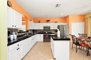 A kitchen with white cabinets, a black countertop, a stainless steel refrigerator, and a dining area with a table and chairs. - 8BR Disney Villa | Pool, W/D, WiFi, Billiards (Kissimmee)