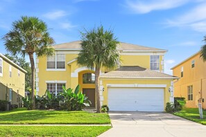 A yellow two-story house with a white garage door, surrounded by well-maintained grass and palm trees. - 8BR Disney Villa | Pool, W/D, WiFi, Billiards (Kissimmee)