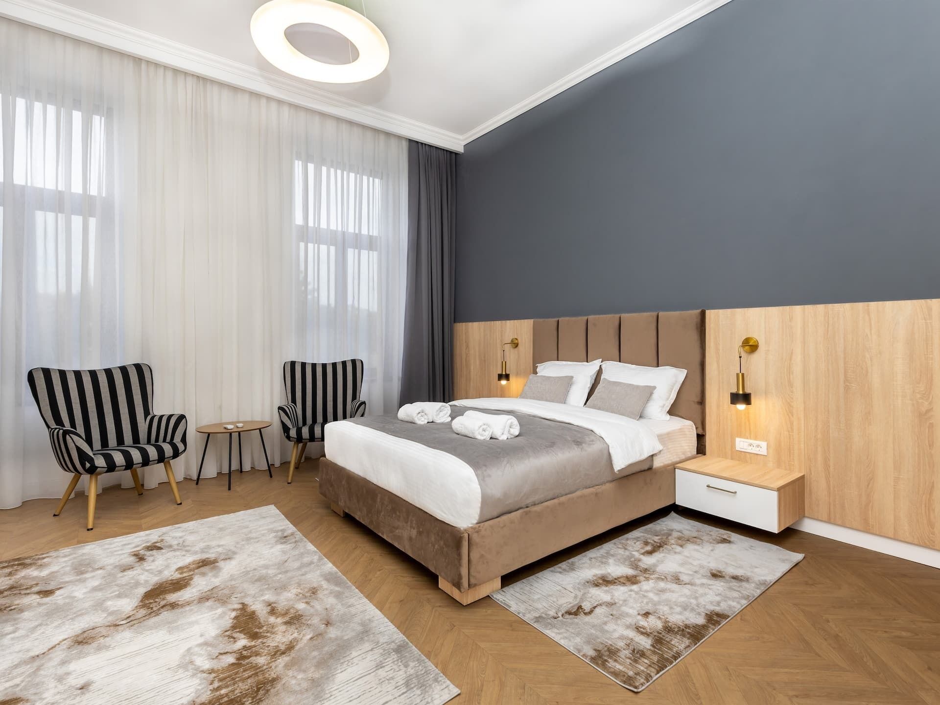 Una habitación de hotel moderna con una cama grande, dos sillones, una mesita y una mesita de noche con lámpara.