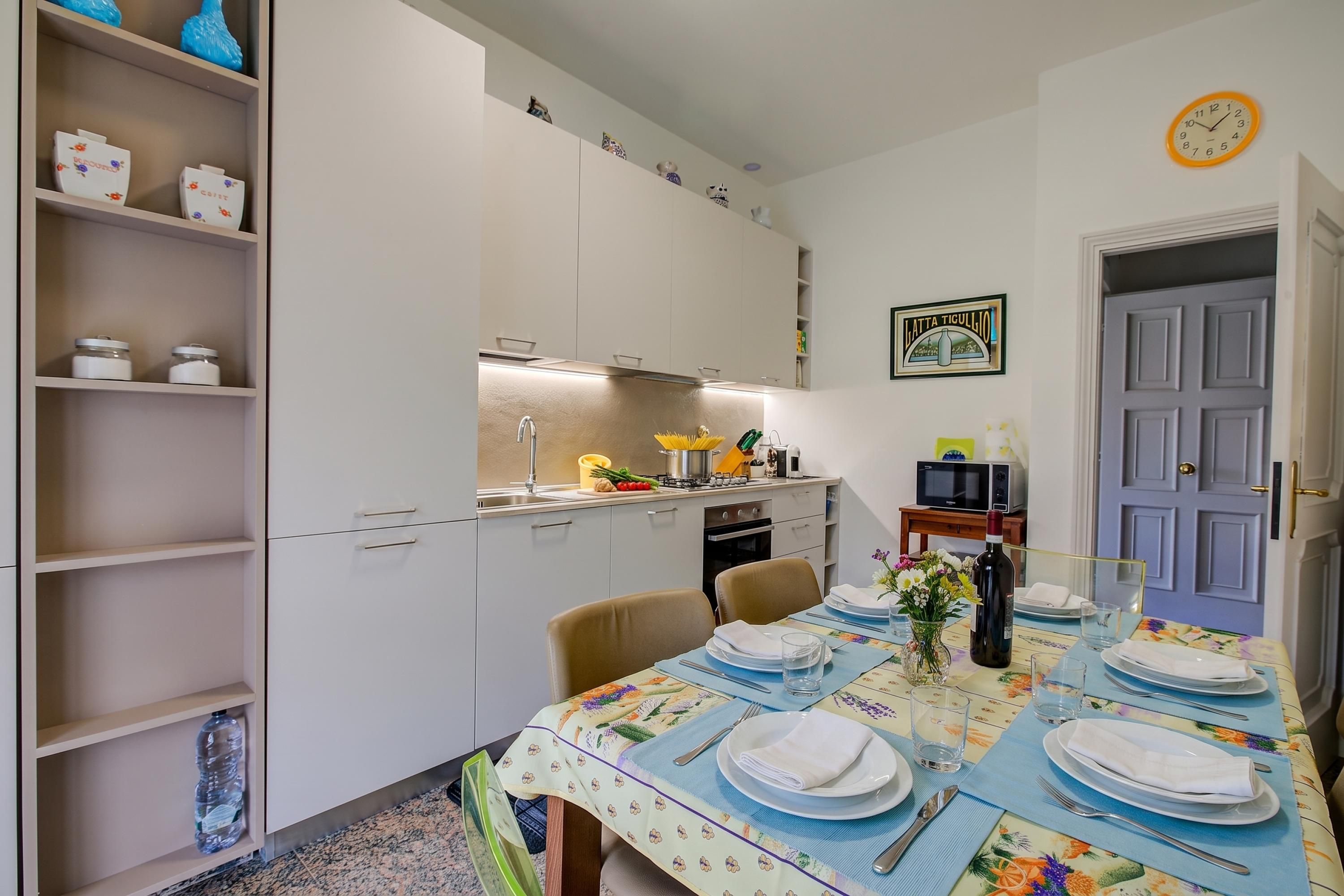 A dining area with a table set for four, a kitchen in the background, and a door with a sign.
