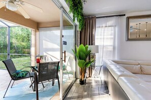 A modern living room with a glass door leading to a balcony, a sofa, a dining table with chairs, and a potted plant. - 3BR Tropical Oasis w/ Pool & Foosball near Disney (Kissimmee)