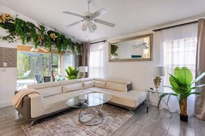 Un salon moderne avec un canapé d’angle, une table basse en verre, des ventilateurs de plafond et de grandes fenêtres avec des rideaux.