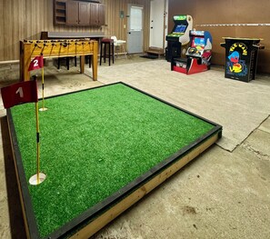 Ein Minigolfplatz mit grünem Kunstrasen, zwei nummerierten Fahnen und einem Holztisch mit einem Schild darüber.