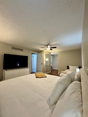 A hotel room with a large bed, a flat-screen TV, a ceiling fan, and a door leading to another room. - Central 5-Bed King Retreat with Hot Tub and Game Room (Indianapolis)