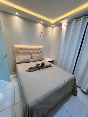 Een moderne slaapkamer met een gestoffeerd hoofdbord, een net opgemaakt bed, een nachtkastje en een gordijn.