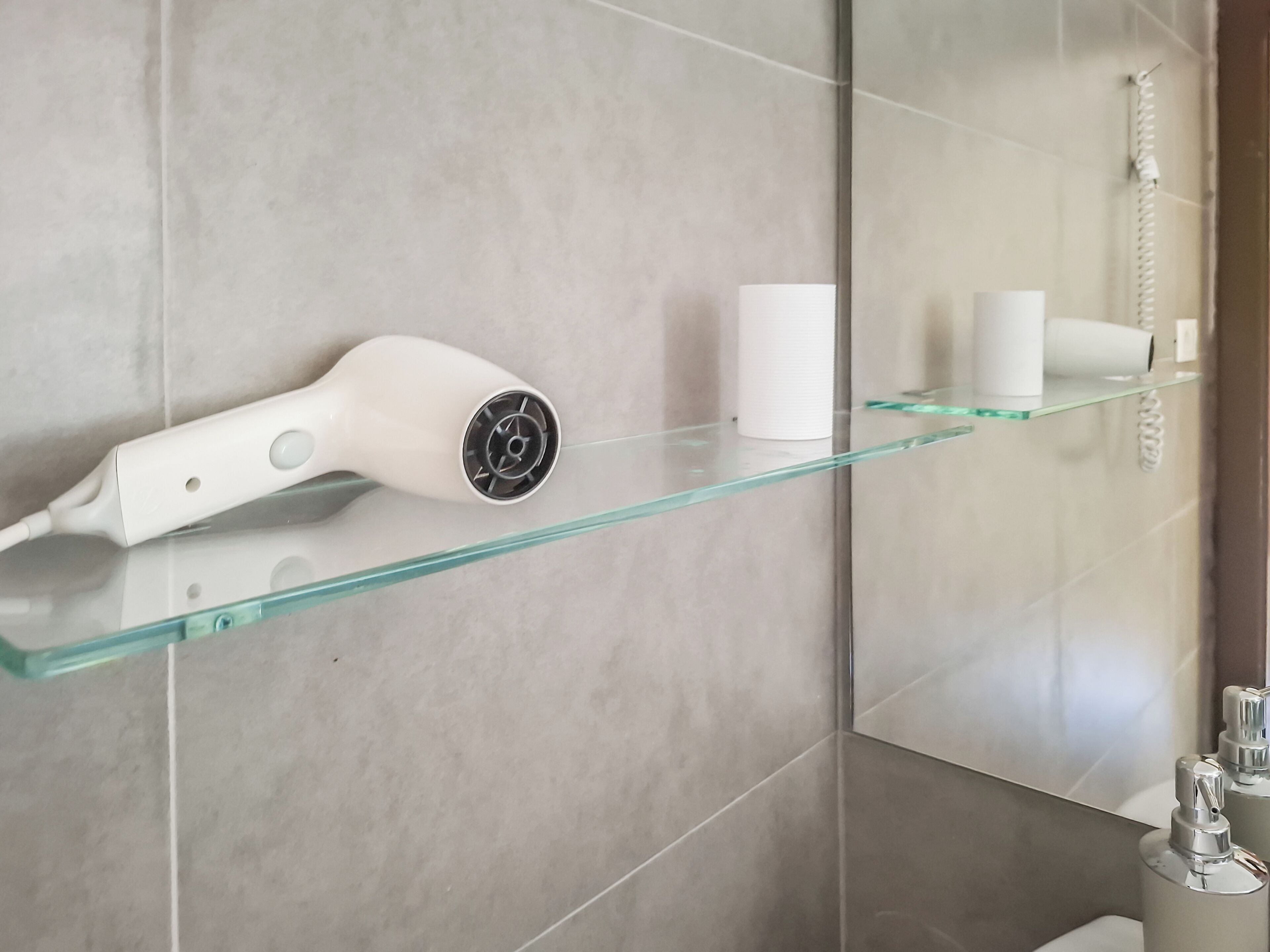 A bathroom with a glass shelf holding a hairdryer and a cup, and a shower area with a hose.