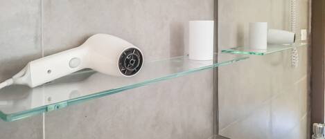 A bathroom with a glass shelf holding a hairdryer and a cup, and a shower area with a hose.