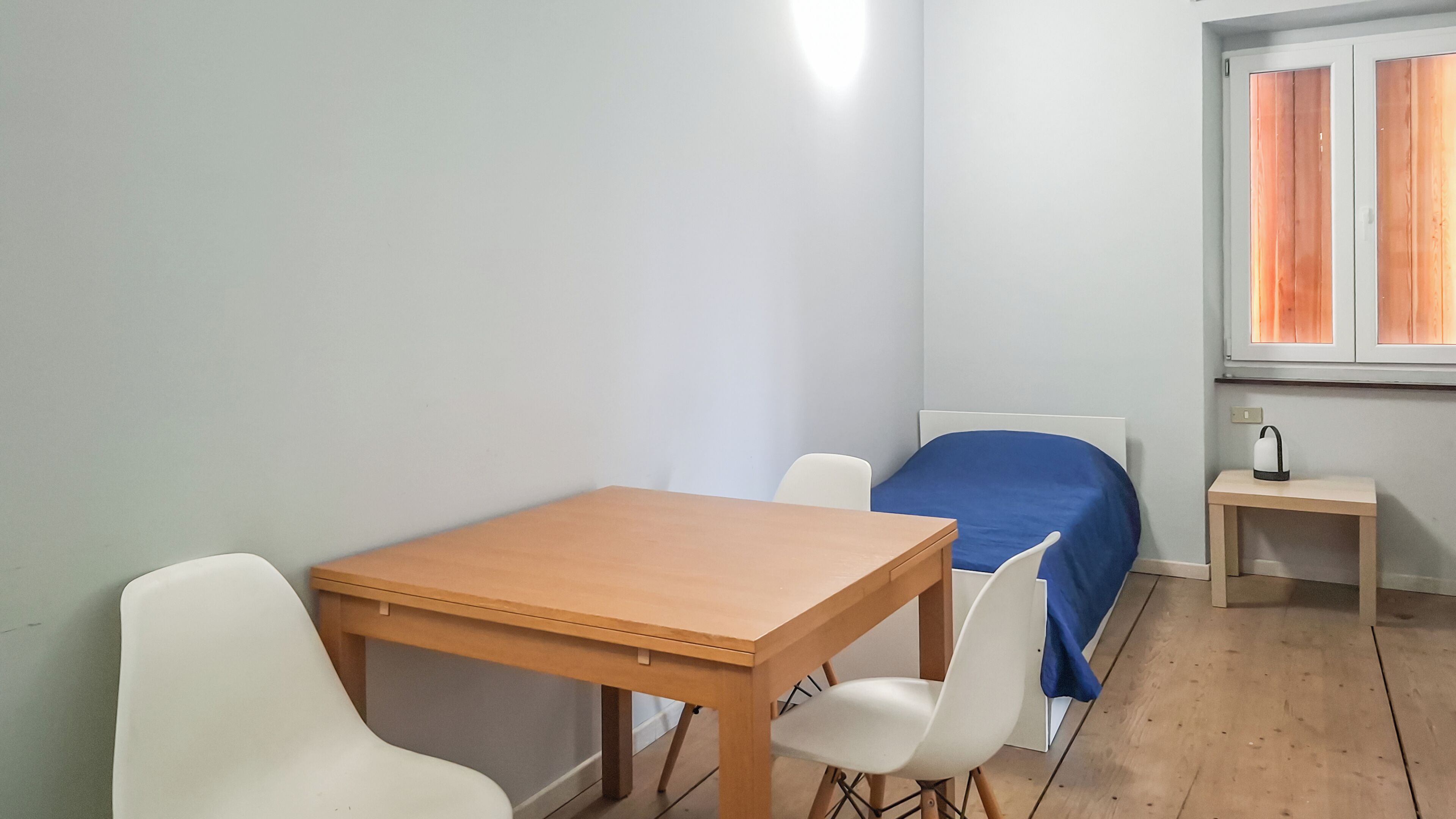 Una habitación pequeña y sencilla con una mesa de madera, dos sillas blancas, una cama individual con cubrecolcha azul y una mesita de noche con una tetera.
