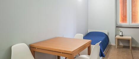 A small, simple room with a wooden table, two white chairs, a single bed with a blue cover, and a small bedside table with a kettle.