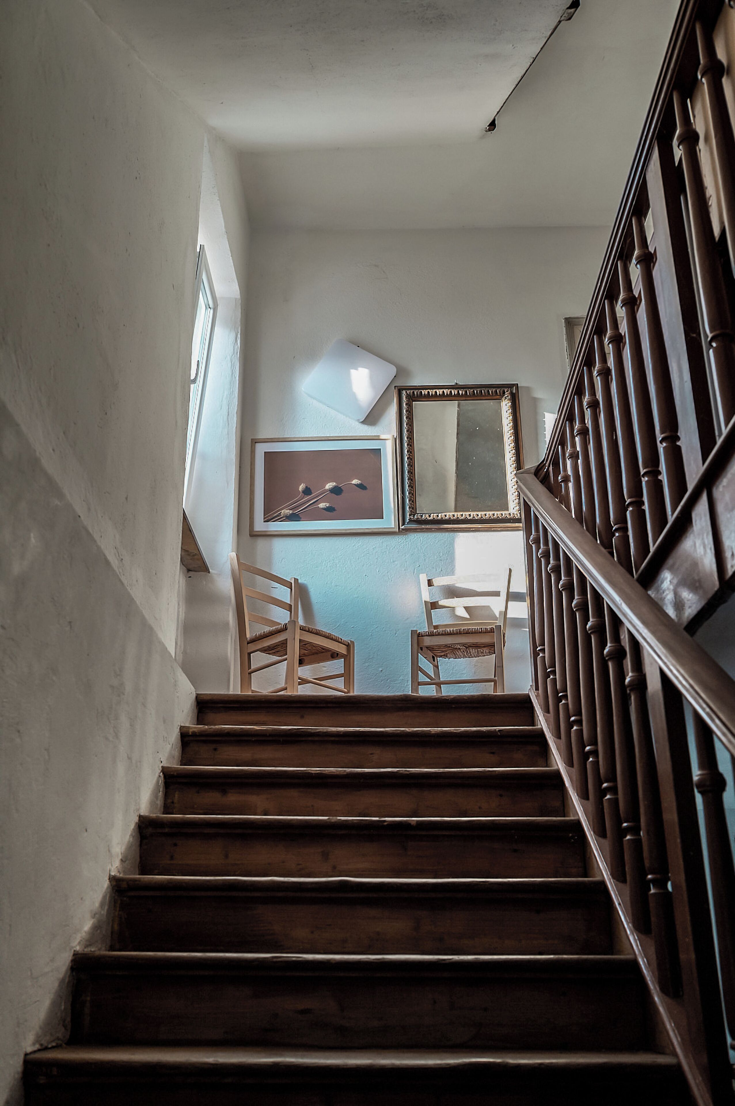 A wooden staircase with a handrail, leading to a landing with two chairs and a framed picture.