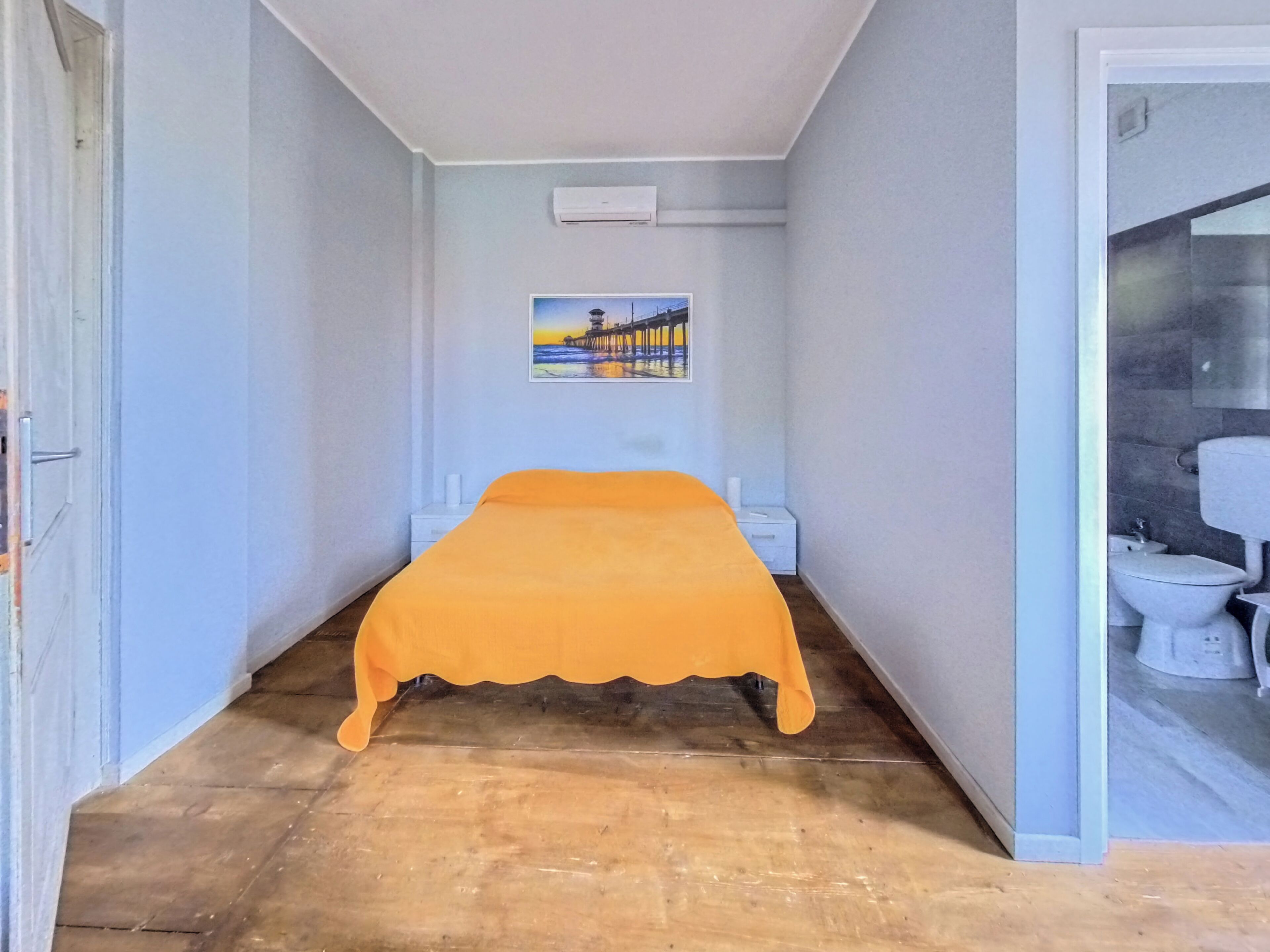 Un pasillo estrecho que conduce a una habitación con una cama cubierta con una manta amarilla, un cuadro en la pared y un baño con un inodoro a la vista.