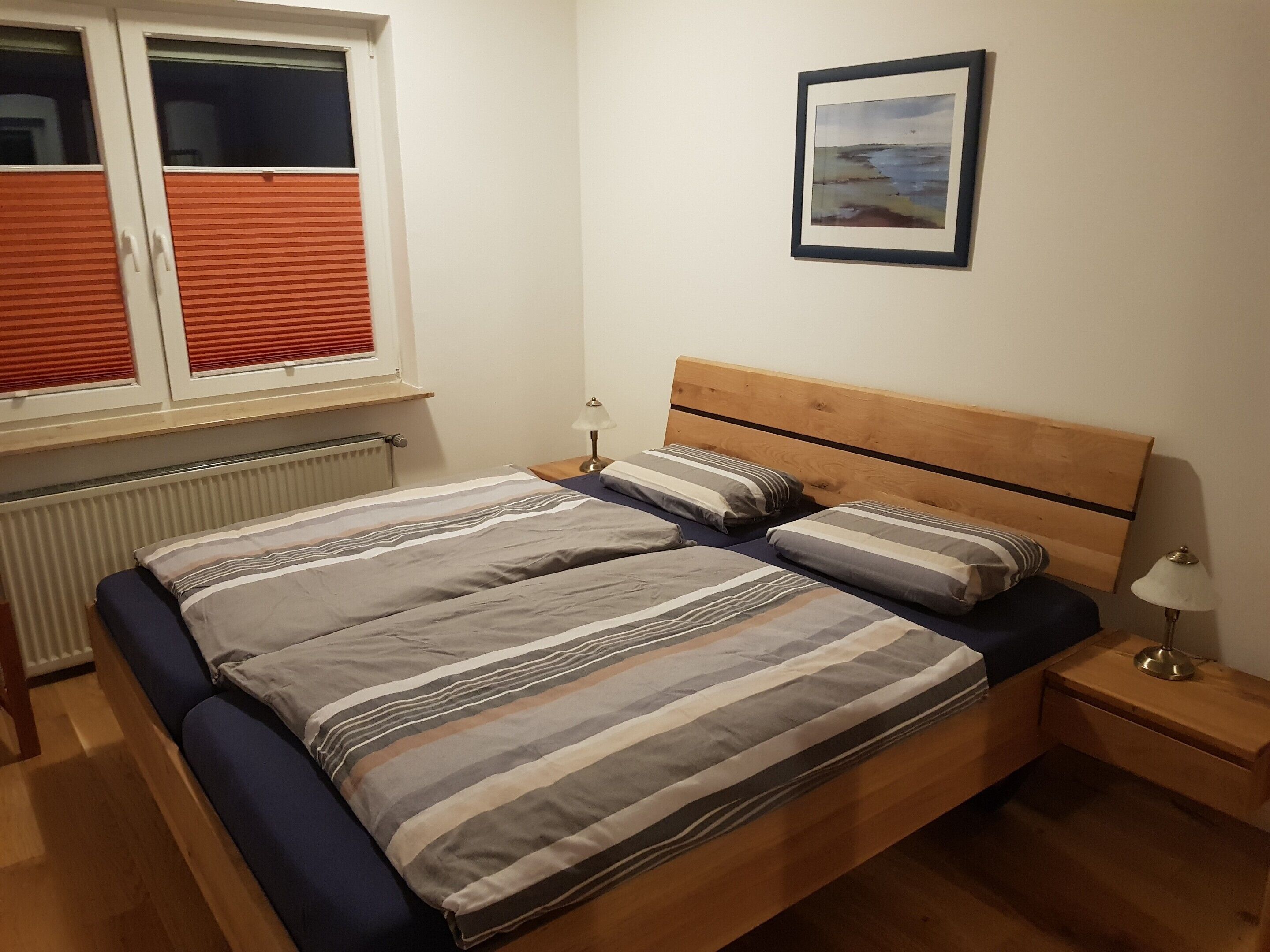 Ein Schlafzimmer mit einem Bett, einem Nachttisch, einer Lampe, einem Fenster mit Jalousien und einem gerahmten Bild an der Wand.