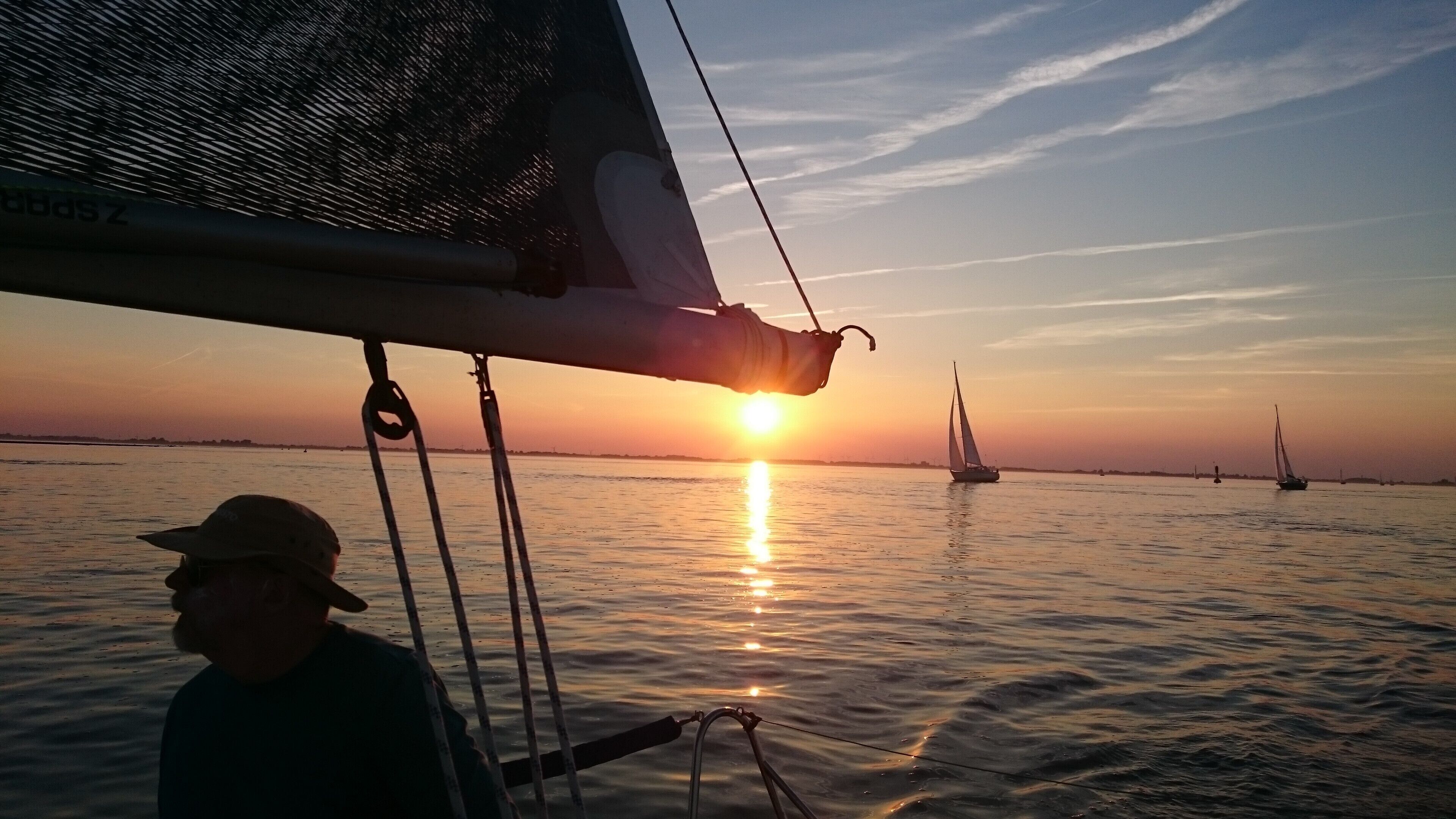 Vista del atardecer desde un bote, con una persona en primer plano y veleros a lo lejos.