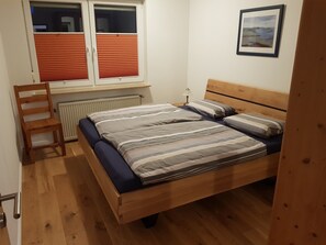 A bedroom with a wooden bed, a chair, a radiator, a window with blinds, and a framed picture on the wall. - Apartment 'Fewo Tenge EG Links' with Covered Terrace, Private Garden and Wi-Fi (Wangerland)