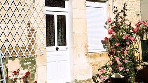 A two-story stone building with a white door and window shutters, a metal fence, and a potted plant with pink flowers. -  grande propriété close (Polisot)