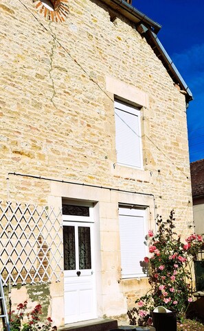 Ein zweistöckiges Steinhaus mit weißer Tür und Fensterläden, einem Metallzaun und einem Topf mit rosa Blüten.