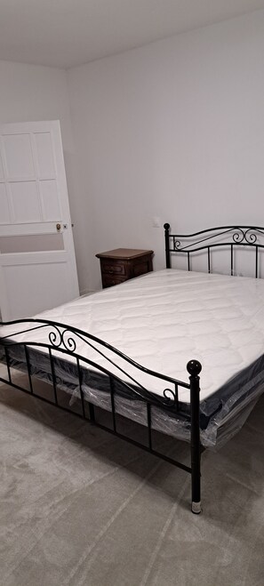 A metal-framed bed with a mattress in a room with a white door and a small wooden nightstand. -  grande propriété close (Polisot)