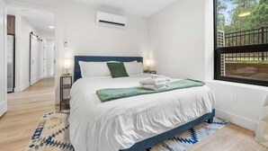 A modern bedroom with a bed, bedside tables, a window with a view, and an air conditioning unit.