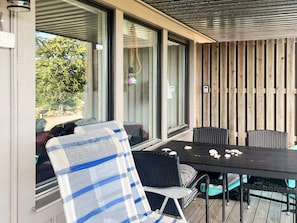 An outdoor patio with a table and chairs, a view of greenery through the windows, and a wooden wall. - 4 Star Holiday Home in Uddevalla (Uddevalla)