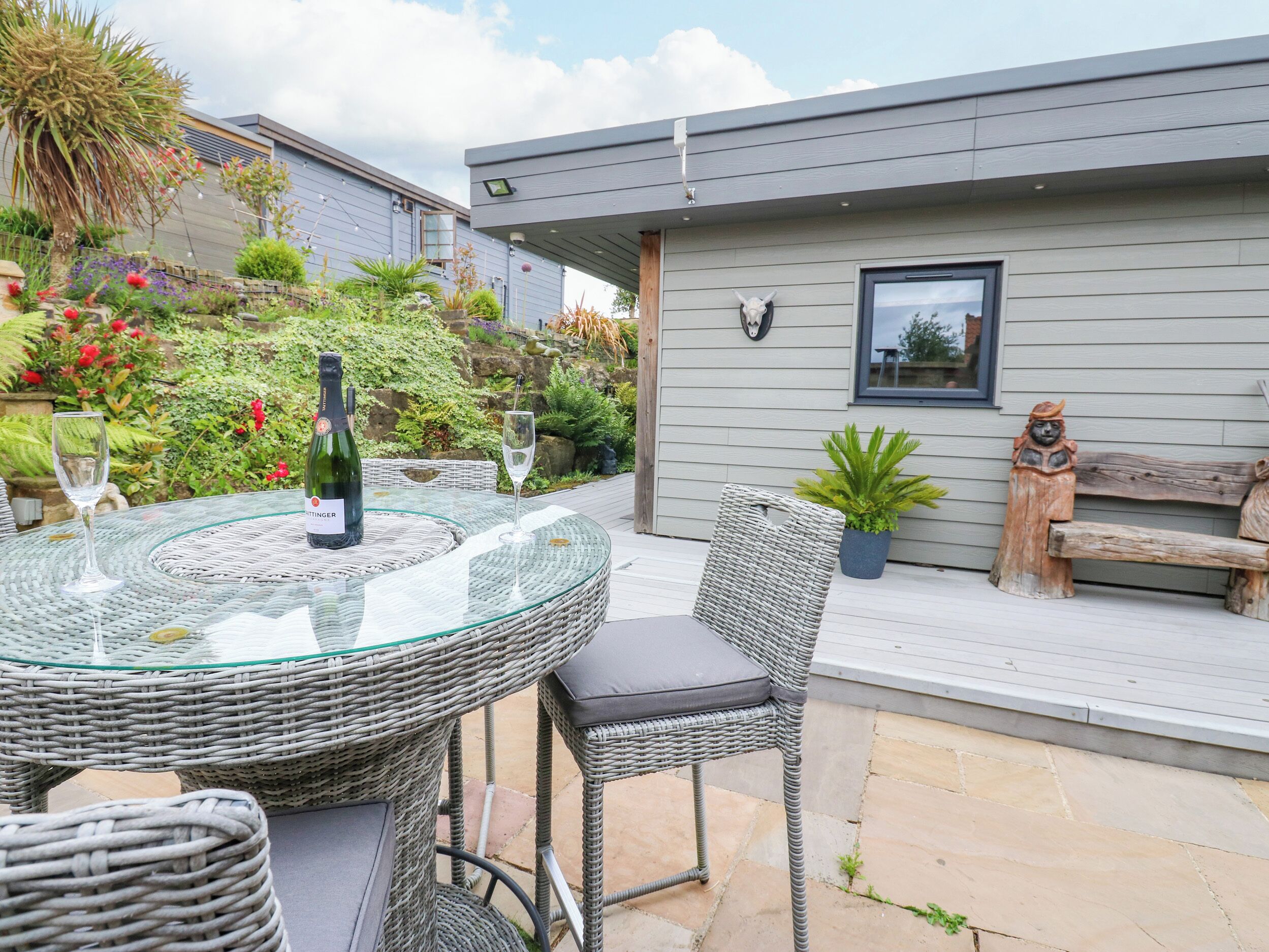 A glass-top table with a bottle and glass, wicker chairs, a wooden bench, and a potted plant.