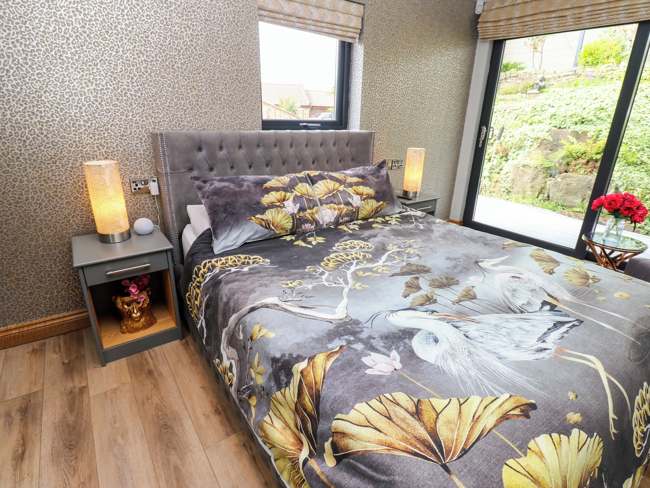 A bedroom with a tufted headboard, a bed with a floral print bedspread, a nightstand with a lamp, and a view of greenery outside.