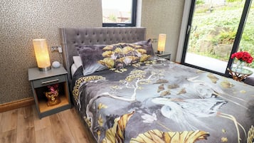 A bedroom with a tufted headboard, a bed with a floral print bedspread, a nightstand with a lamp, and a view of greenery outside.
