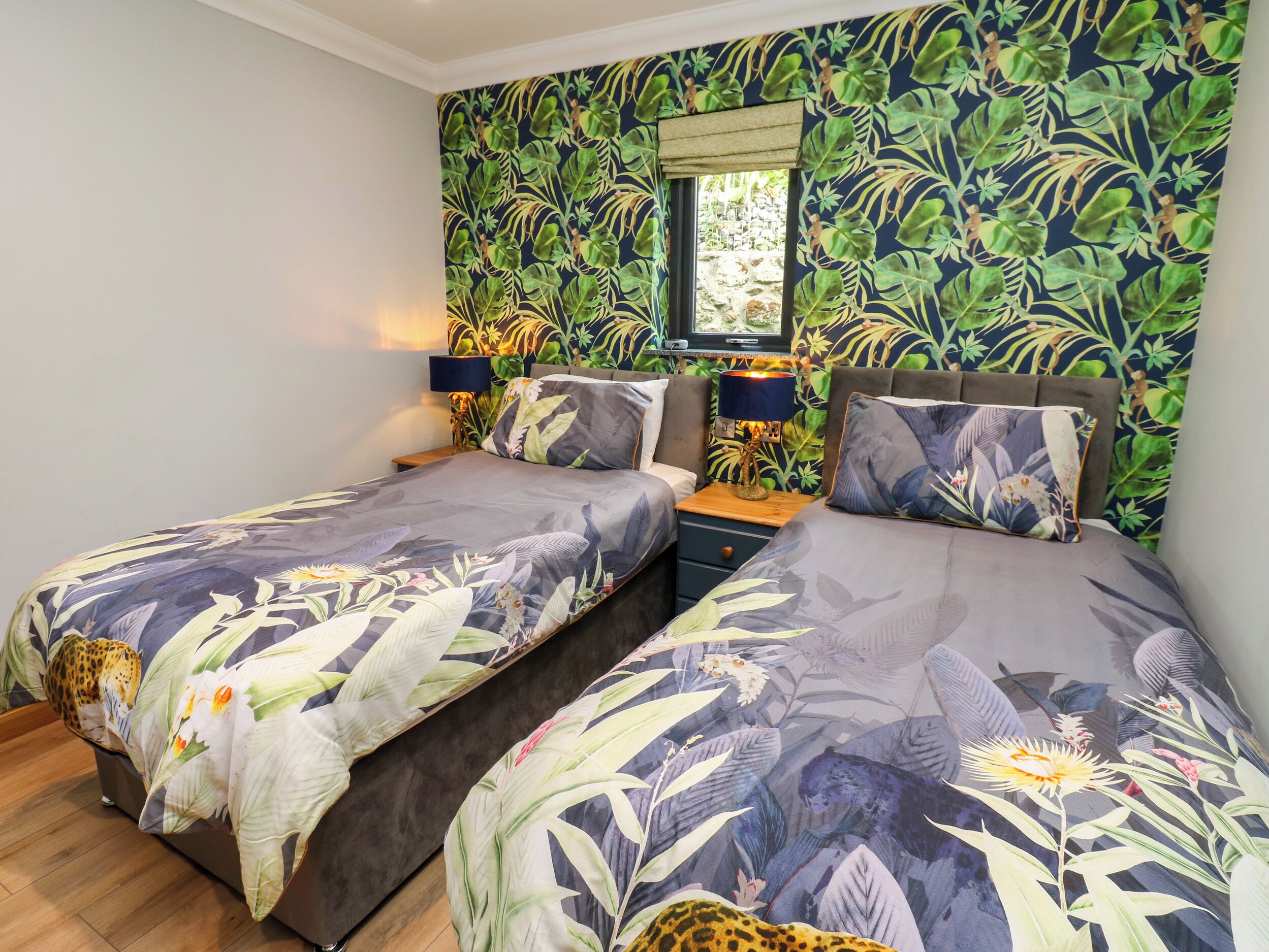 A bedroom with two beds, a nightstand with a lamp, a window with a view, and a tropical wallpaper design.