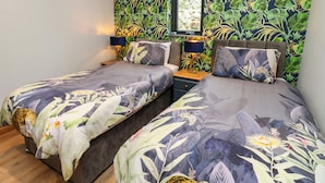 Un dormitorio con dos camas, una mesita de noche con lámpara, una ventana con vistas y un diseño de papel tapiz tropical.