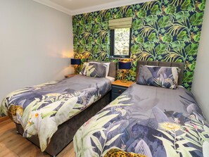 A bedroom with two beds, a nightstand with a lamp, a window with a view, and a tropical wallpaper design. - Caesars Lodge (Darlington)
