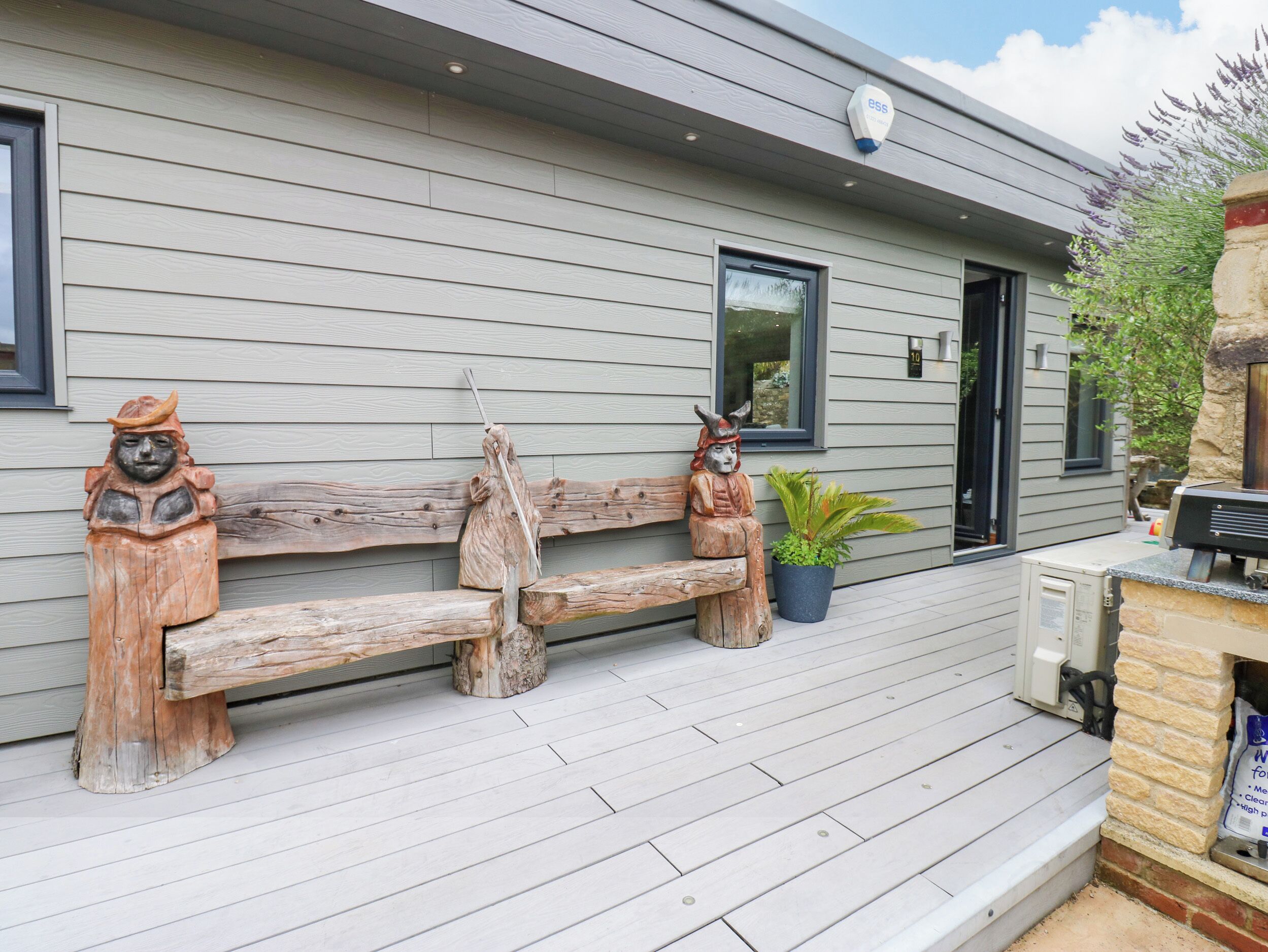 Un banc en bois orné de deux sculptures en bois à chaque extrémité, un pot de fleurs et un espace barbecue sur une terrasse en bois.