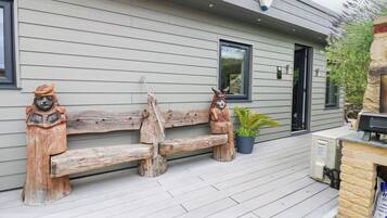 Un banc en bois orné de deux sculptures en bois à chaque extrémité, un pot de fleurs et un espace barbecue sur une terrasse en bois.
