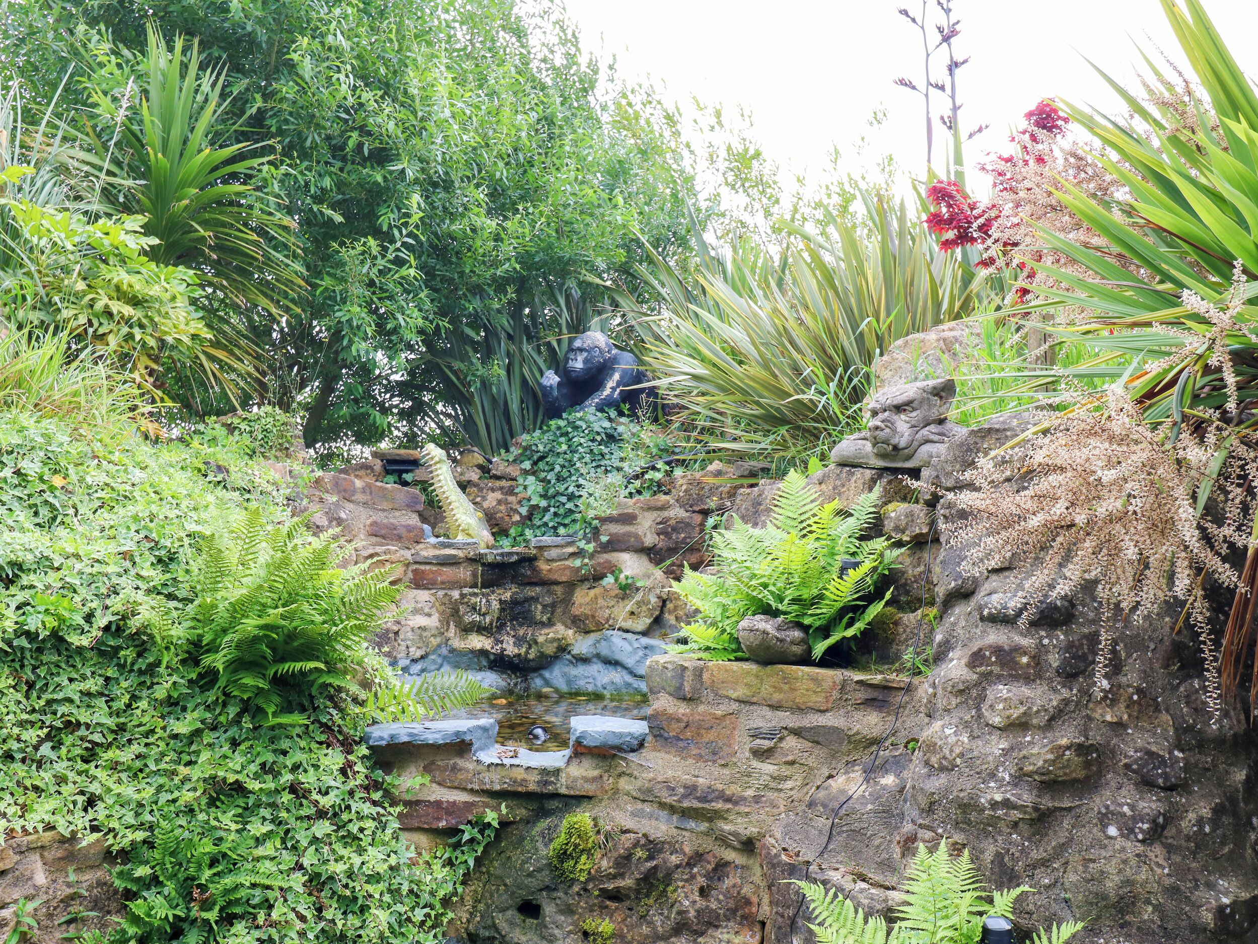 Un jardín con muro de piedra, una fuente pequeña y diversas plantas.