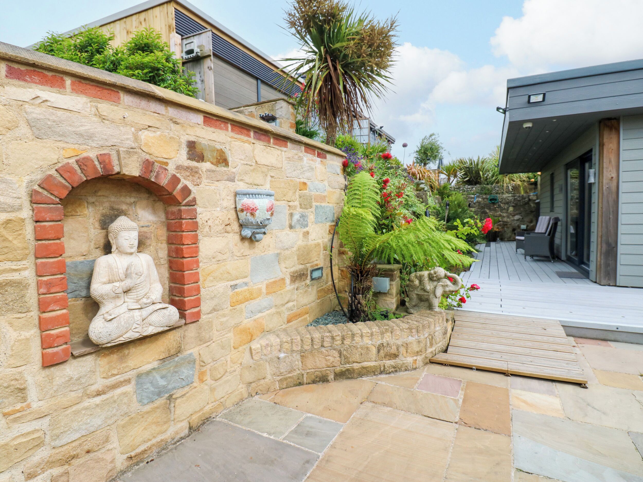 Una estatua de Buda de piedra en un nicho, un muro de piedra con una placa, y una terraza de madera con mobiliario exterior.