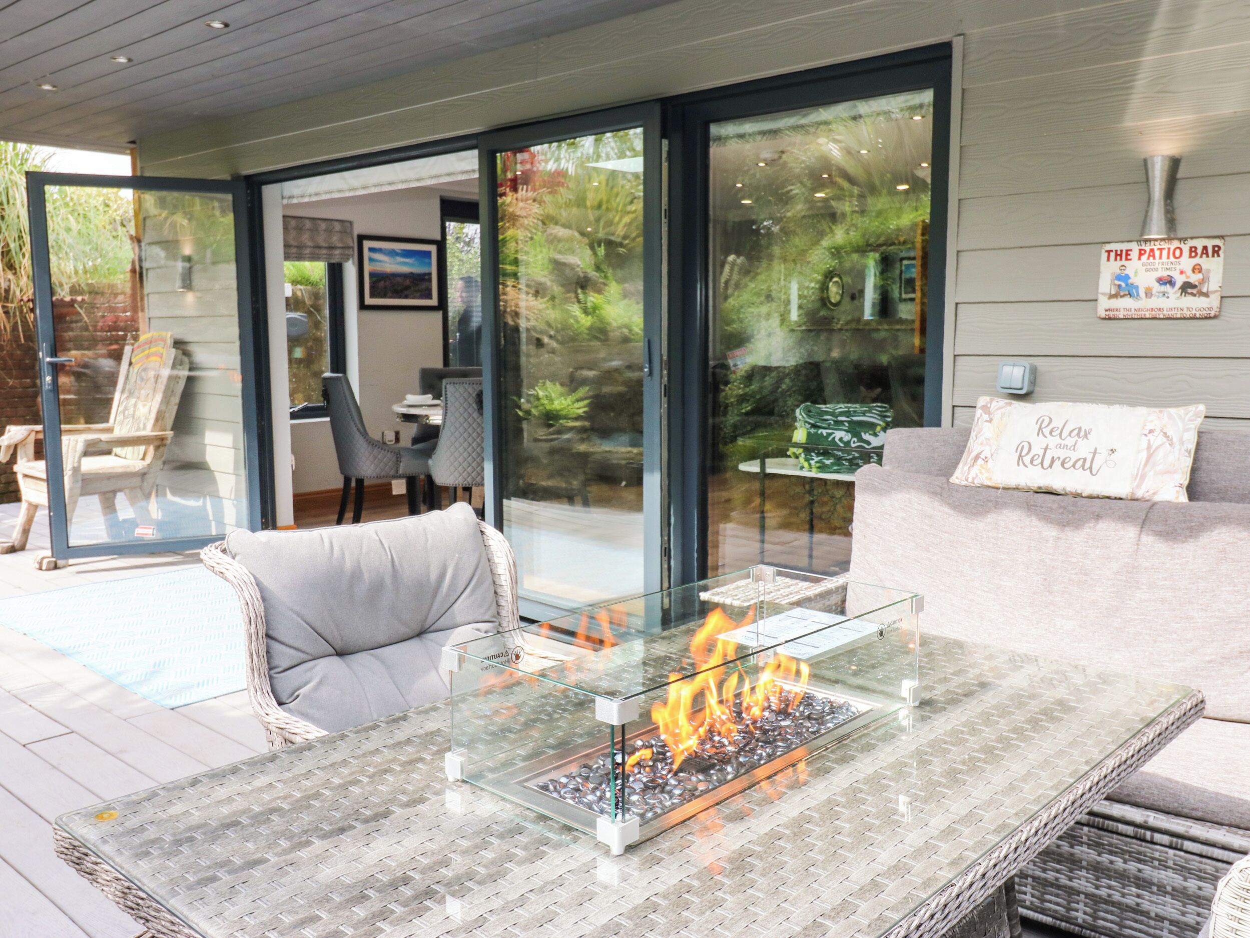 Un espace extérieur moderne aménagé avec des sièges, une table à plateau de verre dotée d'un foyer intégré, des meubles en osier et une vue sur un jardin, accessible par des portes vitrées.