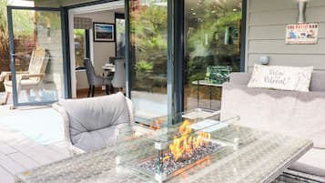 Uma área moderna de estar ao ar livre, com mesa de vidro e lareira embutida, mobiliário de vime e vista para um jardim através de portas de vidro.