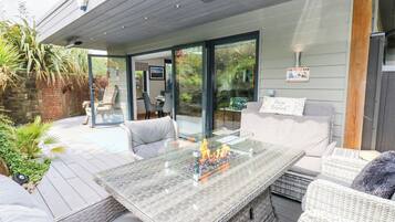 Een terras buiten met een tafel met glazen blad, rieten meubels en een vuurplaats.