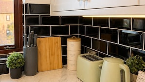 Una cucina con mobili bianchi, zoccolo a parete in piastrelle nere, un tagliere di legno, un portacoltelli, un tostapane, un bollitore e due contenitori etichettati 'tea' e 'coffee'.