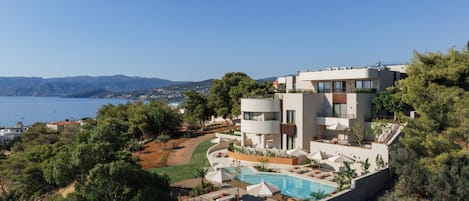 Un hôtel moderne à plusieurs étages, doté d’une piscine, entouré de verdure et offrant une vue sur un plan d’eau.