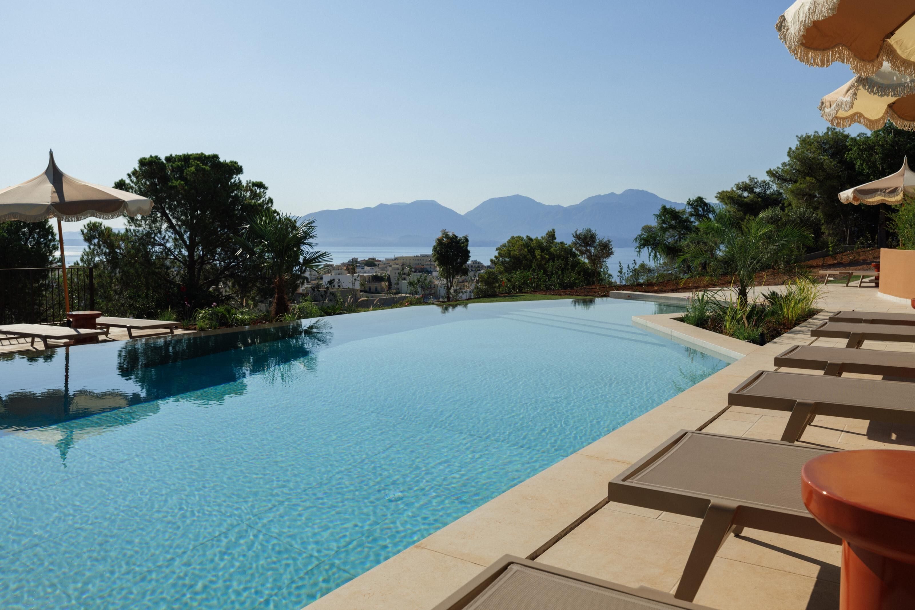 A clear view of an infinity pool with lounge chairs and umbrellas, surrounded by greenery and mountains in the background.