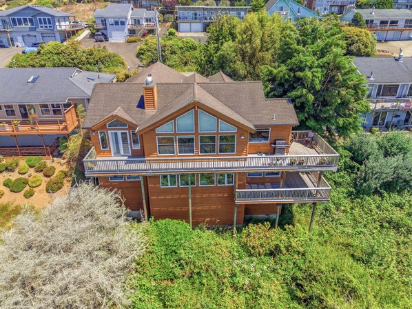 A two-story wooden house with a balcony, surrounded by greenery and other houses. - Panoramic Ocean Views, Grill, Wraparound Decks (Pacific City)