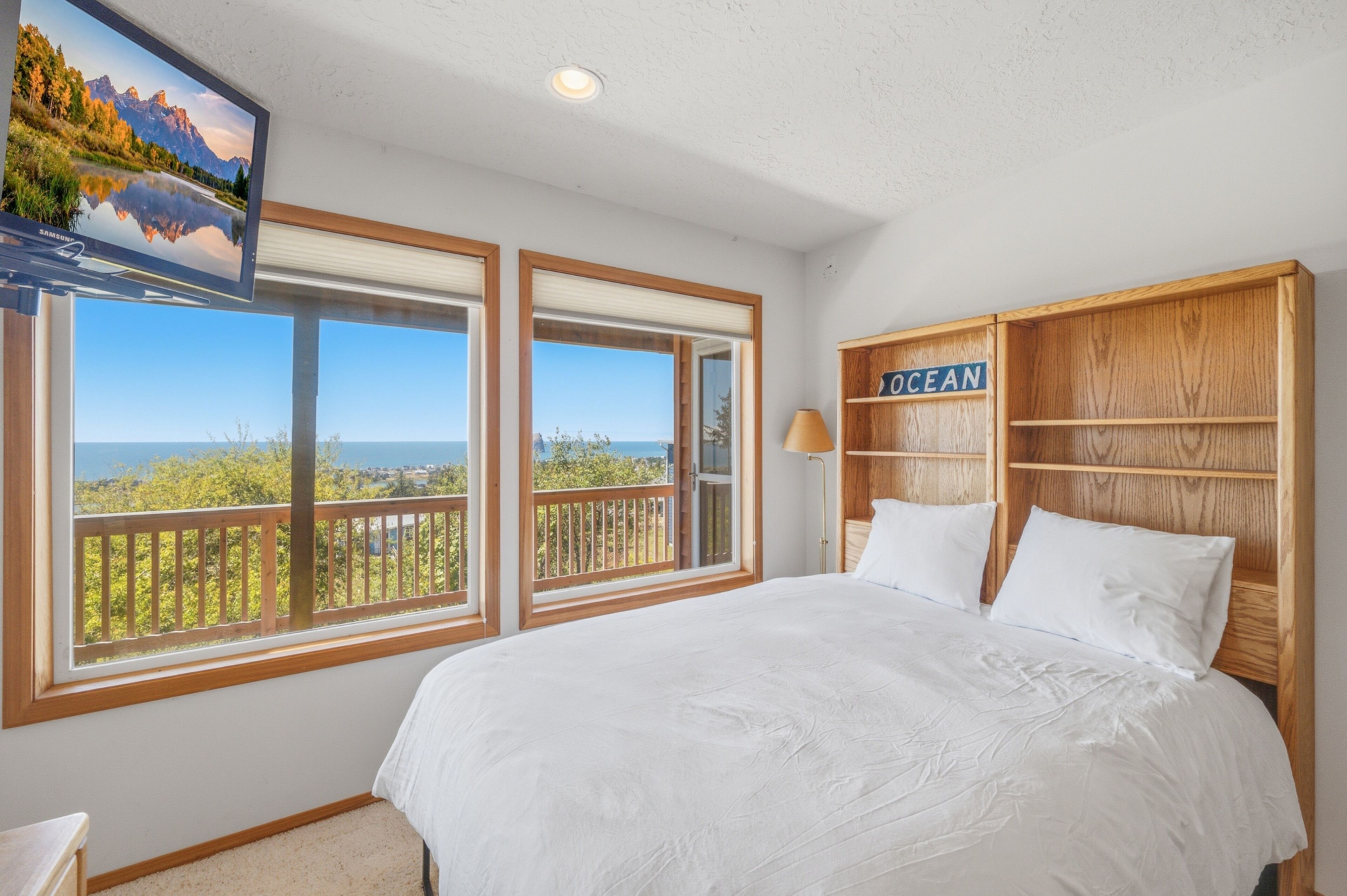 A bedroom with a large bed, two windows with ocean views, a TV on the wall, and a wooden shelf labeled 'OCEAN'.