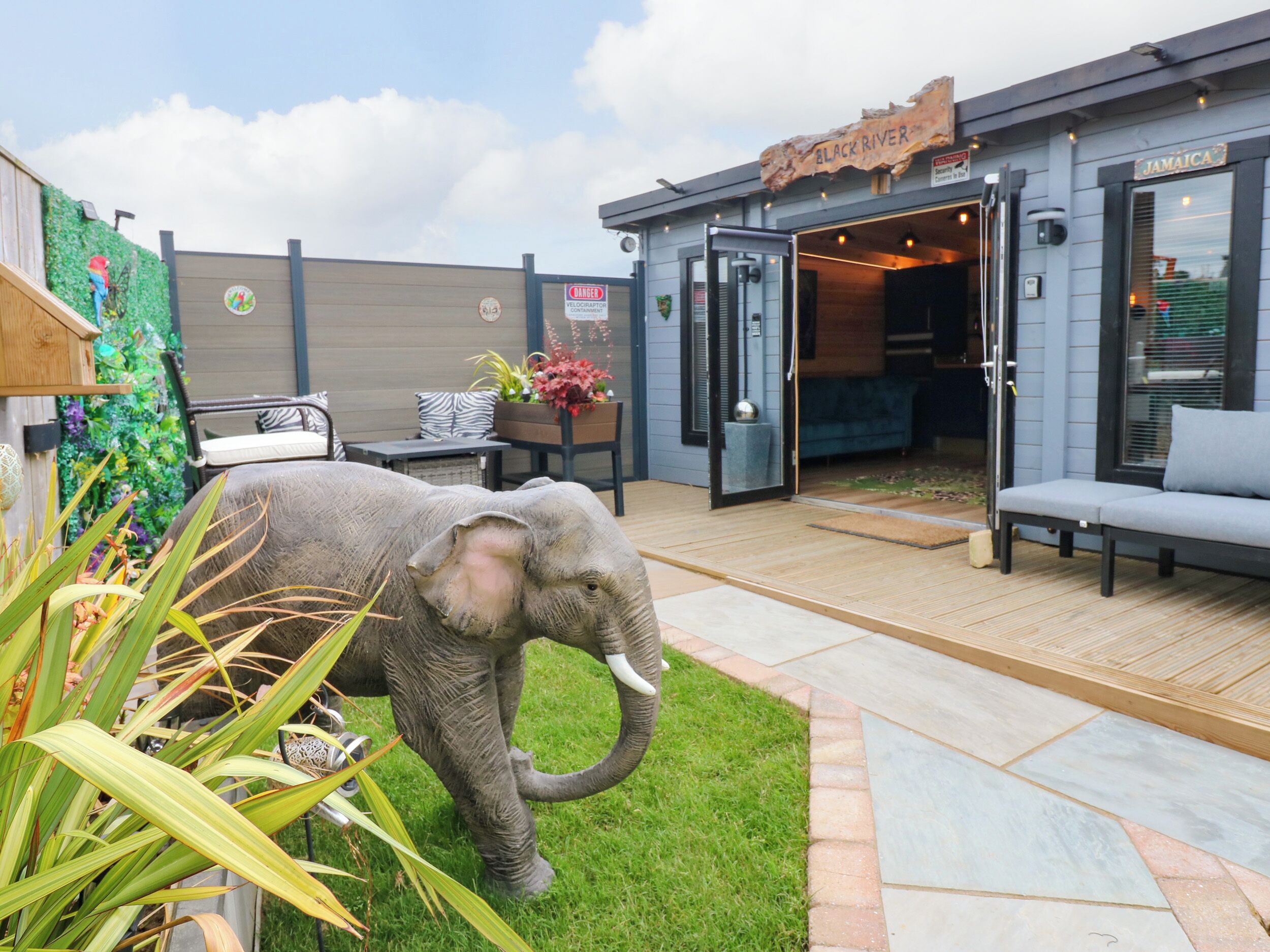 Una estatua de elefante en un jardín con un muelle de madera, una zona de estar con un sofá y sillones, y un edificio con un cartel que dice 'BLACK RIVER'.