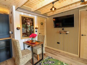 A room with a wooden ceiling, a television mounted on the wall, a mirror with a framed picture, wicker chairs, a small table with a vase of flowers, and a leopard print rug.